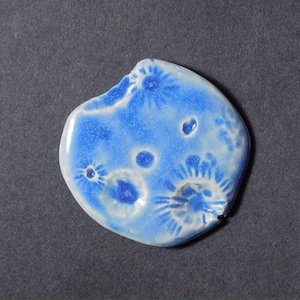 May include: A blue and white ceramic tile with a unique, organic shape and a textured surface. The tile features a design of white flowers and dots on a blue background.