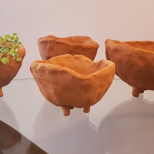 May include: Four terracotta pots with three legs each. The pots are unglazed and have a rough, textured surface. One pot has a small green plant inside.