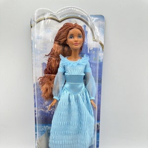 May include: A plastic-packaged doll of Ariel from Disney's The Little Mermaid. The doll is wearing a light blue dress with a white lace trim. She has long, brown hair and brown eyes. The packaging includes the Disney logo and the words "The Little Mermaid" and "Ariel on Land".