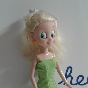 May include: A doll with blonde hair, blue eyes, and a green dress with a floral skirt. The doll is wearing pink shoes and small orange earrings. A white hair accessory is on top of the doll's head.