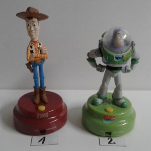 Puede incluir: Dos figuras de personajes de la película de animación Toy Story. Woody, un vaquero, está sobre una base roja. Buzz Lightyear, un astronauta, está sobre una base verde. Ambas bases tienen un botón y el texto "ON SENSOR".