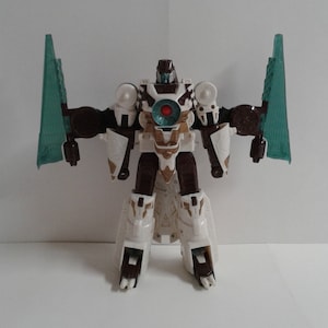 May include: A white, brown, and teal robot toy with wings. The robot has a red eye and a white and gold chest piece.
