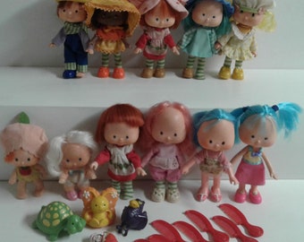 May include: A collection of vintage Strawberry Shortcake dolls, including Strawberry Shortcake, Orange Blossom, Raspberry Tart, Lemon Meringue, Blueberry Muffin, and others. The dolls are dressed in their signature outfits and have bright, colorful hair. There are also accessories such as a turtle, a butterfly, and combs.