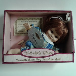May include: A porcelain doll with brown hair and a blue and white plaid dress. The doll is sitting in a pink box with the words "Collector's Choice Poseable Bean Bag Porcelain Doll" on the front.