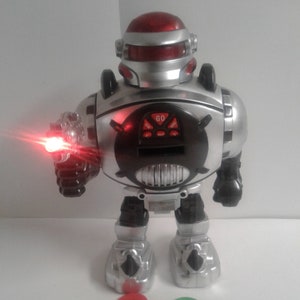 Op de afbeelding: Een zilveren en zwarte robot speelgoed met een rood licht op zijn arm. De robot heeft een zwart gezicht met een digitaal display waarop "GO" staat. De robot staat op een wit oppervlak met vijf kleurrijke ringen.