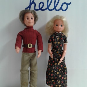 May include: Two vintage dolls, one with brown hair wearing a red sweater, brown pants, and brown shoes, and the other with blonde hair wearing a floral dress and brown sandals.