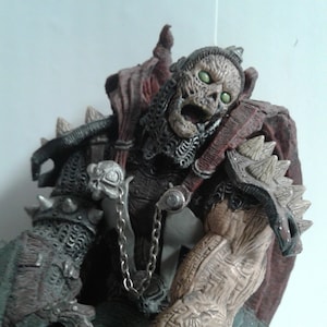 May include: A detailed action figure of a skeletal warrior with a red cape and a chain. The figure has a green and brown colour scheme and is posed in a dynamic fighting stance.