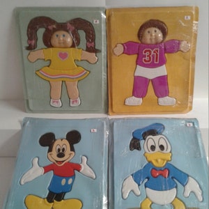May include: Four plastic puzzle pieces, each individually packaged. The puzzles depict a Cabbage Patch Kid girl wearing a yellow dress, a Cabbage Patch Kid boy wearing a pink shirt with the number 31 on it, Mickey Mouse, and Donald Duck.