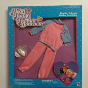 May include: A pink and blue jogging outfit for dolls. The outfit includes a long-sleeved sweatshirt with blue sleeves and pink trousers. The outfit is packaged in a blue box with a heart-shaped window. The box also features the text "The Heart Family" and "La Famille Boncoeur".