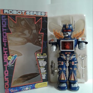 Pode incluir: Um brinquedo robô azul e dourado com uma tela iluminada e efeitos sonoros. O robô está segurando uma arma prateada e está em pé sobre uma base de plástico branco. A caixa diz "Robot Series Sound-Light-Motion" e "S.L.M. Robot".