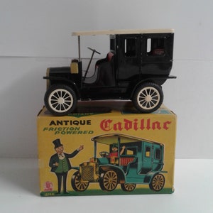 Könnte beinhalten: Schwarzes Spielzeugauto Cadillac mit weißem Dach und einem Reibungsmechanismus. Das Auto befindet sich in seiner Originalverpackung mit einem Vintage-Design. Die Schachtel trägt die Aufschrift "Antique Friction Powered Cadillac" und "Japan".