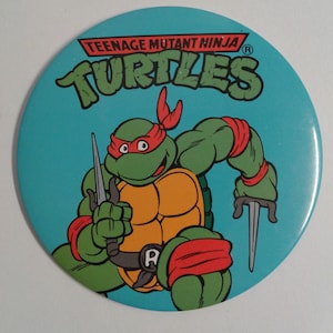 May include: A blue and green Teenage Mutant Ninja Turtles button featuring a cartoon image of Leonardo, one of the four ninja turtles, holding two katanas. The button has the text "Teenage Mutant Ninja Turtles" and a registered trademark symbol.