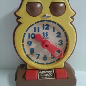 May include: A vintage yellow and brown owl-shaped Answer Clock by Tomy. The clock face has blue numbers and a red clock hand. The base has two red buttons. The word "hello" is written in blue cursive above the clock.