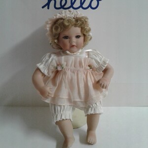 May include: A porcelain doll with blonde curly hair wearing a pink dress and white bloomers. The doll is sitting on a white surface with the word "hello" written in blue above it.