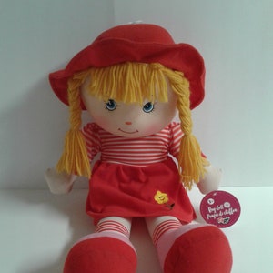 May include: A red and white striped dress with a yellow flower on the front. The doll has blonde yarn hair in pigtails and is wearing a red hat. The doll is sitting on a white surface. The tag says "Rag doll Puppe de chiffon"