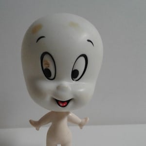 May include: A white plastic figurine of a cartoon ghost with a smiling face and large eyes. The ghost has a simple body with arms outstretched and legs.
