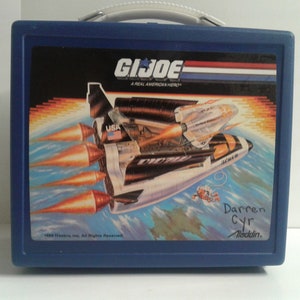May include: Blue plastic lunchbox with a G.I. Joe logo and a space shuttle graphic. The lunchbox has a handle and the text "A Real American Hero" is printed on the front. The lunchbox has a copyright date of 1989.