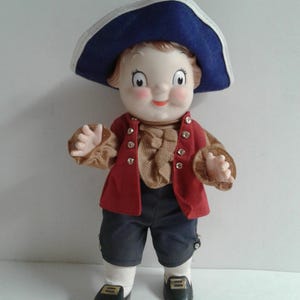 Pu&ograve; includere: Bambola vintage vestita con un abito in stile coloniale. La bambola ha un viso dipinto, indossa un cappello blu con bordo bianco, un gilet rosso e pantaloncini blu scuro. La bambola indossa scarpe nere con fibbie dorate.