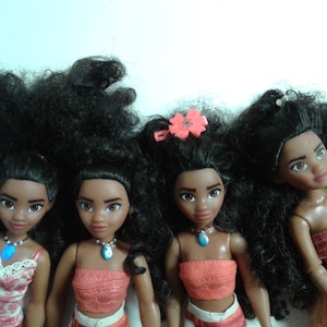 May include: Four Moana dolls with long black curly hair. The dolls are wearing different outfits, including a floral top, a coral top, and a red top. The dolls have brown skin and dark eyes.