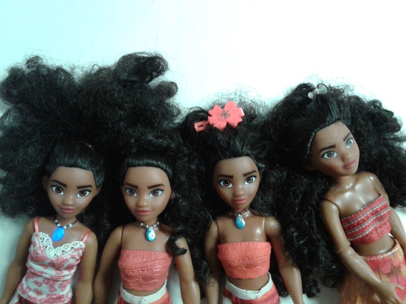 May include: Four Moana dolls with long black curly hair. The dolls are wearing different outfits, including a floral top, a coral top, and a red top. The dolls have brown skin and dark eyes.