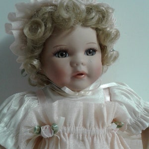 May include: A porcelain doll with blonde curly hair wearing a pink dress and a pink bonnet. The doll has blue eyes and a sweet expression.