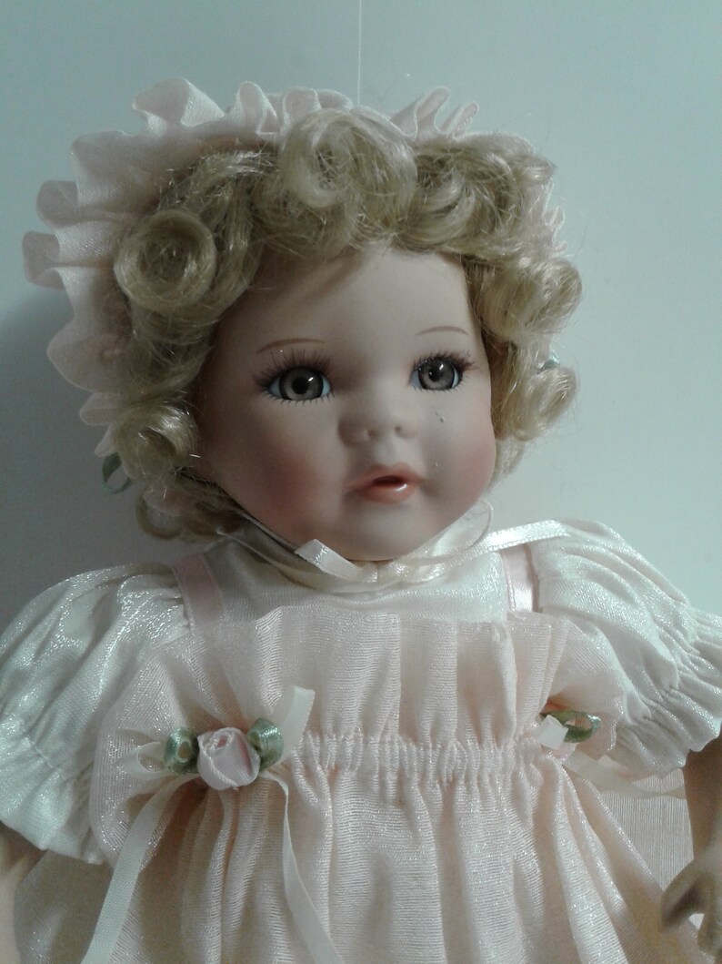 May include: A porcelain doll with blonde curly hair wearing a pink dress and a pink bonnet. The doll has blue eyes and a sweet expression.