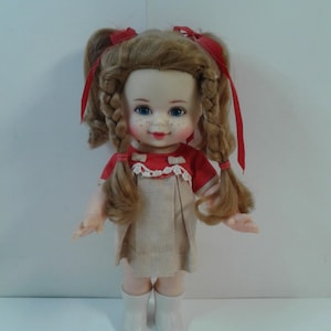 Puede incluir: Una muñeca vintage con coletas rubias trenzadas atadas con cintas rojas. La muñeca viste un vestido rojo y beige y botas blancas. La muñeca tiene ojos azules y una sonrisa pintada, un juguete clásico.