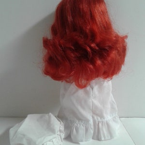 K&ouml;nnte beinhalten: Eine Puppe mit roten Haaren, die ein wei&szlig;es Kleid mit R&uuml;schen am Saum und eine wei&szlig;e Haube tr&auml;gt. Die Puppe steht auf einer wei&szlig;en Oberfl&auml;che.
