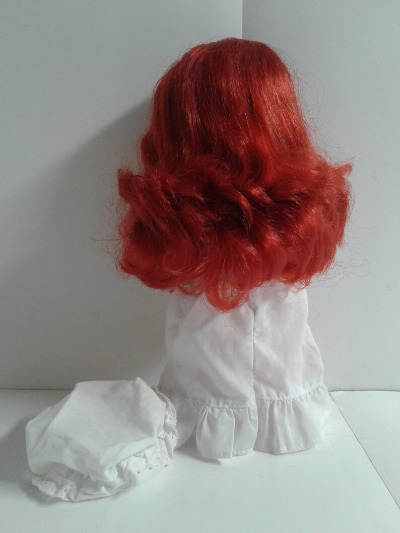 K&ouml;nnte beinhalten: Eine Puppe mit roten Haaren, die ein wei&szlig;es Kleid mit R&uuml;schen am Saum und eine wei&szlig;e Haube tr&auml;gt. Die Puppe steht auf einer wei&szlig;en Oberfl&auml;che.