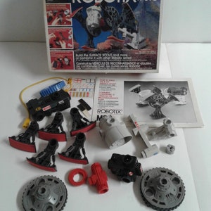 May include: A set of black and grey plastic toy robot parts, including wheels, gears, and arms. The parts are from the Robotix R-310 series, a motorised modular construction system. The box shows a robot with red, black, and grey parts.
