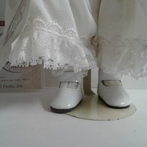 Pode incluir: Boneca de porcelana branca vestindo um vestido branco com detalhes em renda e sapatos brancos. A boneca est&aacute; em p&eacute; em um suporte branco.