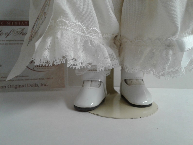 Pode incluir: Boneca de porcelana branca vestindo um vestido branco com detalhes em renda e sapatos brancos. A boneca est&aacute; em p&eacute; em um suporte branco.