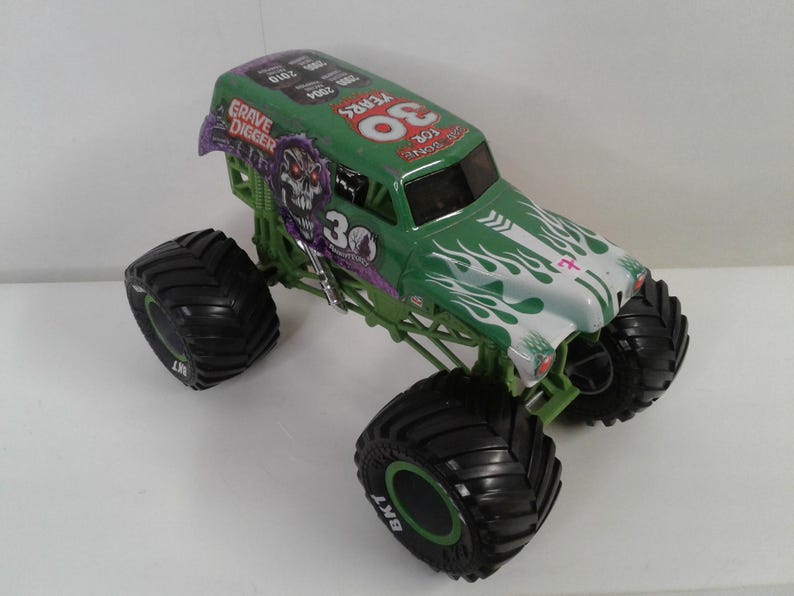 May include: A green and white Grave Digger monster truck toy with large black tyres. The truck features the number "30" and the words "Grave Digger" and "30 Years" on the body. The tyres are marked with "BKT".