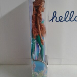 May include: A mermaid doll with long brown hair, wearing a green and blue tail. The doll is packaged in a clear plastic box with a blue and green background.