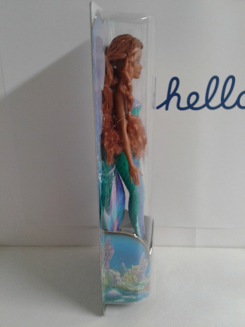 May include: A mermaid doll with long brown hair, wearing a green and blue tail. The doll is packaged in a clear plastic box with a blue and green background.