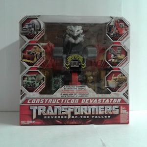 May include: A Transformers Constructicon Devastator toy box featuring a red and gray robot with a large head. The box has a picture of the robot in the center and images of the six vehicles that combine to form the robot around the edges. The box also has the text "Transformers Revenge of the Fallen" on it.