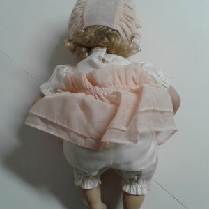 May include: A porcelain doll wearing a pink dress with a white underskirt and a pink bonnet. The doll has blonde hair and is posed sitting with its legs crossed.