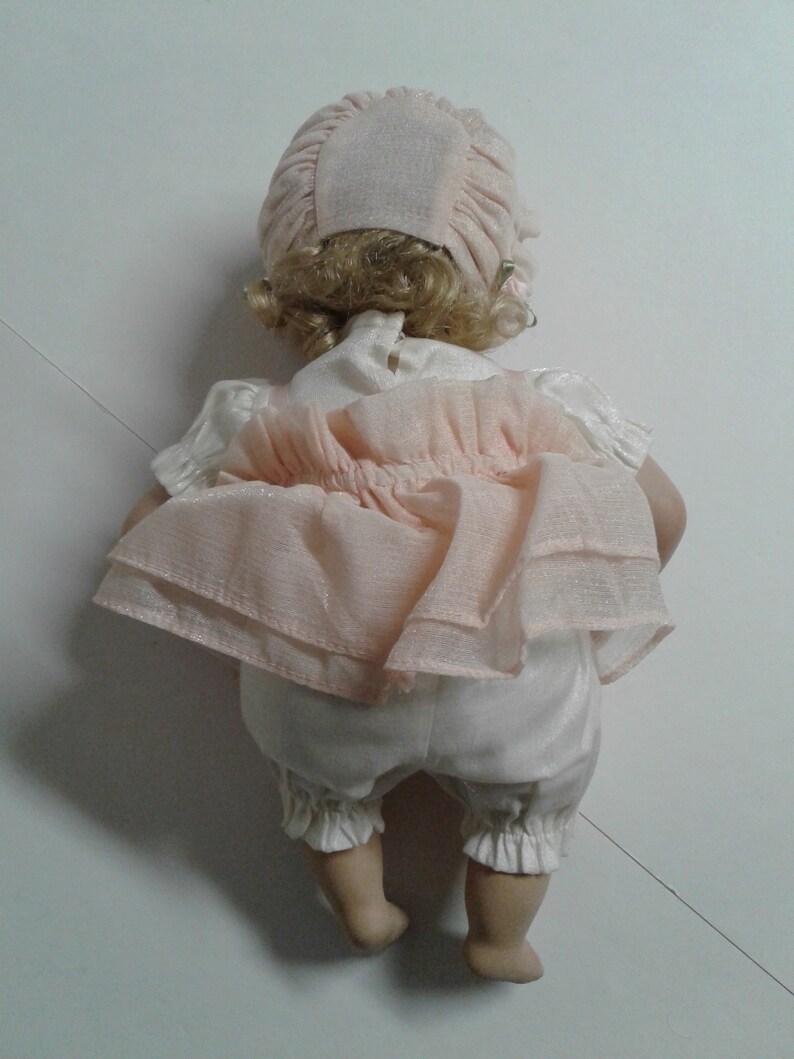 May include: A porcelain doll wearing a pink dress with a white underskirt and a pink bonnet. The doll has blonde hair and is posed sitting with its legs crossed.
