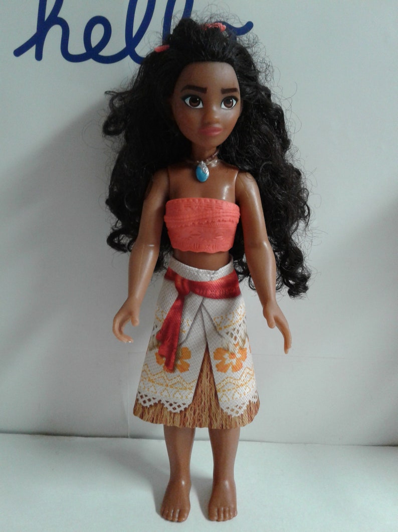 May include: A brown-skinned doll with long black curly hair wearing a coral-colored halter top, a white and brown skirt with floral designs, and a blue necklace.