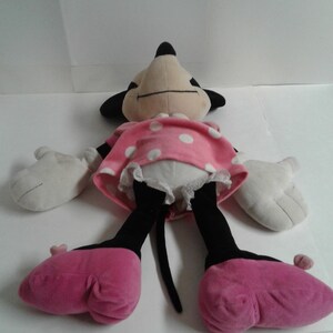 May include: A plush toy of Minnie Mouse, lying on its back with its arms outstretched. The toy is wearing a pink dress with white polka dots and pink shoes. The toy has a black nose and eyes, and a long black tail.