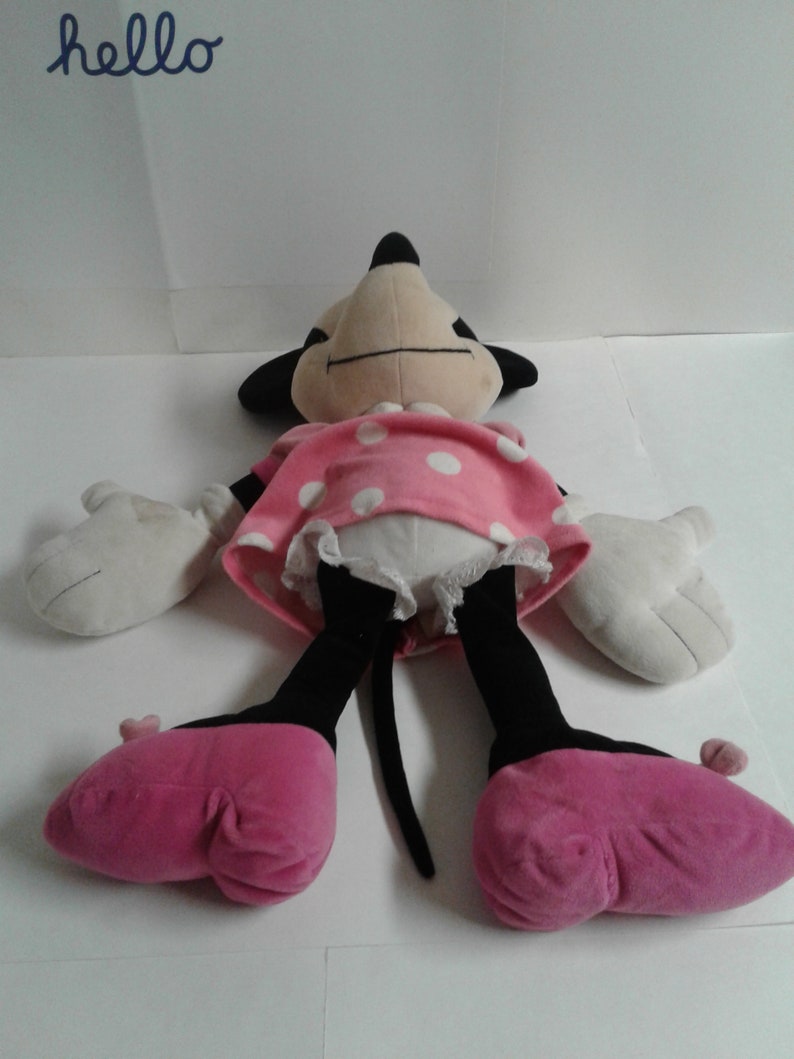 May include: A plush toy of Minnie Mouse, lying on its back with its arms outstretched. The toy is wearing a pink dress with white polka dots and pink shoes. The toy has a black nose and eyes, and a long black tail.