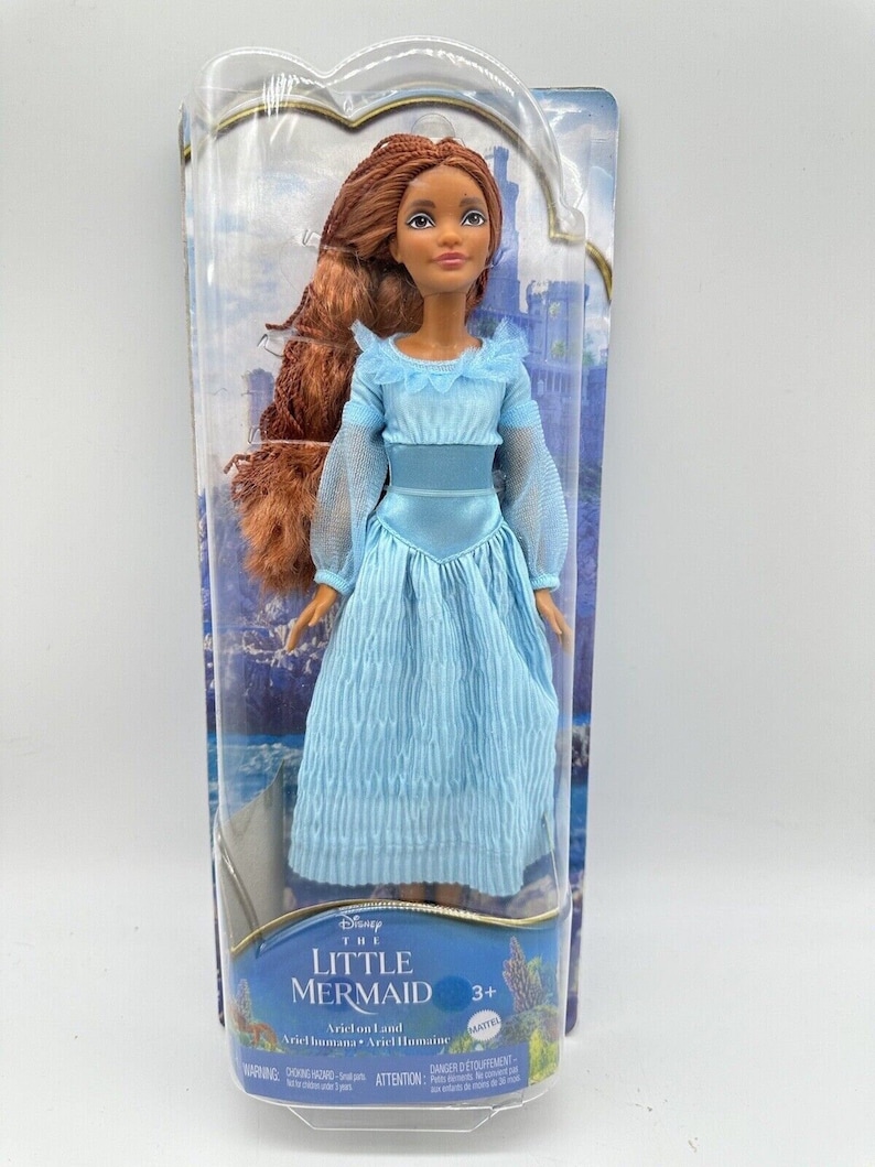 May include: A plastic-packaged doll of Ariel from Disney's The Little Mermaid. The doll is wearing a light blue dress with a white lace trim. She has long, brown hair and brown eyes. The packaging includes the Disney logo and the words "The Little Mermaid" and "Ariel on Land".