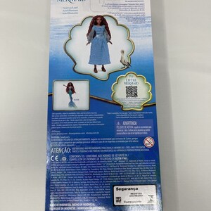 May include: A blue and white doll of Ariel from The Little Mermaid wearing a blue dress and holding a seashell. The doll is standing on a white background with a blue and white background behind her. The packaging includes the text "The Little Mermaid" and "Ariel on Land".
