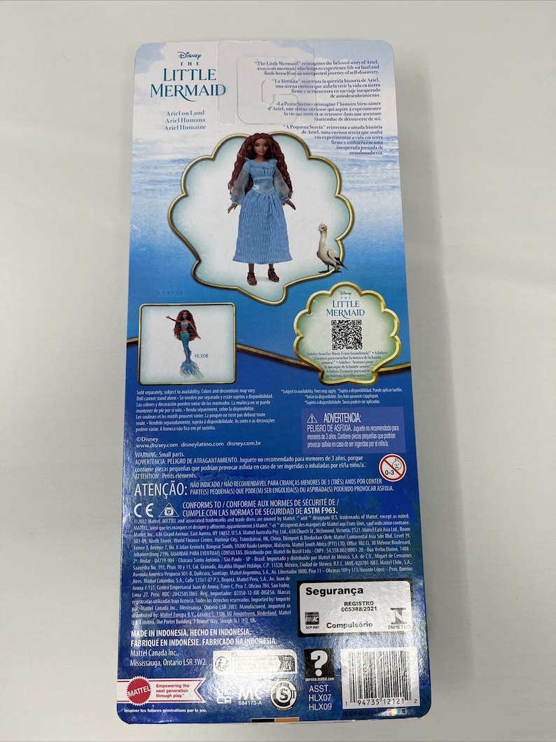 May include: A blue and white doll of Ariel from The Little Mermaid wearing a blue dress and holding a seashell. The doll is standing on a white background with a blue and white background behind her. The packaging includes the text "The Little Mermaid" and "Ariel on Land".