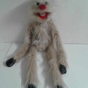 May include: A handmade puppet with a shaggy, light brown and white fur body. It has a white, fluffy head, blue eyes, a red nose, and a red mouth. The puppet's feet are black. This whimsical puppet is designed for entertainment.