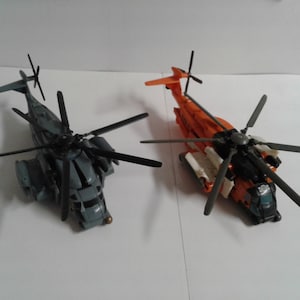 Puede incluir: Dos helicópteros de juguete, uno gris y otro naranja, con rotores negros. El helicóptero gris tiene una cabina detallada y un rotor de cola negro. El helicóptero naranja tiene un esquema de pintura blanco y negro y un rotor de cola negro.