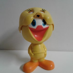 Pode incluir: Um brinquedo de plástico amarelo do Piu-Piu dos Looney Tunes. O brinquedo tem uma cabeça removível que revela um rosto branco com olhos azuis e uma boca vermelha. O brinquedo está em pé sobre uma superfície branca.