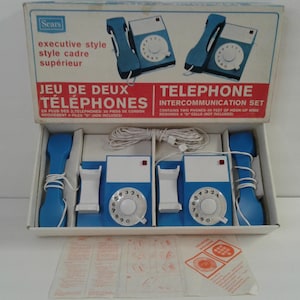 Peut inclure: Ensemble d'interphone téléphonique Sears vintage dans sa boîte d'origine. L'ensemble comprend deux téléphones bleus et blancs avec des cadrans rotatifs, des combinés et un fil de connexion. La boîte contient du texte en anglais et en français.