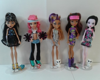 May include: Five Monster High dolls are shown, each with distinct outfits and accessories. The dolls have different hair colours and styles, and are posed on clear stands. One doll has pink hair and pom-poms, another has a black dress.