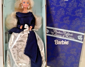 May include: A blonde Barbie doll in a navy blue velvet bodice and a metallic silver skirt with a floral pattern, wearing white earrings and a pearl necklace. The doll is inside a box labelled An Avon Exclusive Winter Velvet Barbie.